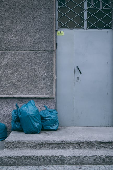 The image shows a set of four blue rubbish bags placed on a concrete step in front of a grey industrial-style door with a black handle, situated beside a textured concrete wall. Above the door, there is a partly visible metal security grille covering a window or vent, indicating an urban environment. The bags appear to be filled with waste and are made of durable plastic with slightly wrinkled surfaces. The concrete surface of the step and the surrounding pavement are clean, with minimal signs of wear. A small yellow sticker or sign is affixed to the wall near the top of the door. This scene likely depicts an area used for private waste disposal or collection outside a commercial or residential building, where external rubbish bags are temporarily stored before collection by waste removal services such as Waste Removal Hackney. The overall setting emphasizes a typical site for independent rubbish management or on-site clearance in the context of non-local-authority waste handling.