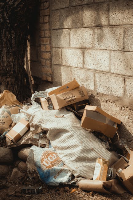 A pile of mixed waste materials and cardboard boxes is gathered against a rough concrete wall in an outdoor area, with some items partially contained within paper or plastic packaging. The waste includes flattened cardboard cartons, some with visible printed labels, alongside crumpled paper and plastic bags, with a large fabric sack partially covering the debris. The background features a weathered brick wall and a portion of a tree trunk on the left side. The scene appears to capture an instance of rubbish awaiting collection or removal, with natural daylight illuminating the scene. This setting reflects a typical example of waste that might require private rubbish removal services in Hackney, managed by companies such as Waste Removal Hackney, especially when access for regular collection is limited or difficult, as suggested by the surrounding environment.