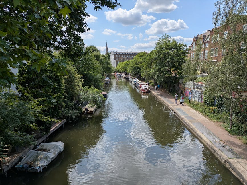 A view of a narrow urban canal lined with lush green trees on both sides, with the water surface reflecting a partly cloudy sky. On the right side, a paved walkway runs parallel to the canal, featuring a few pedestrians walking along it. Several parked cars are visible along the street beyond the trees, and a graffiti-covered wall is partially obscured by foliage. In the background, a mixture of modern and historic buildings, including a prominent church steeple, rise above the trees under a mostly sunny sky. The scene is peaceful and typical of city-centre waterside areas, illustrating an environment where private waste management or on-site clearance may be relevant for maintaining surrounding spaces, as implied by the context of waste removal services in Hackney.