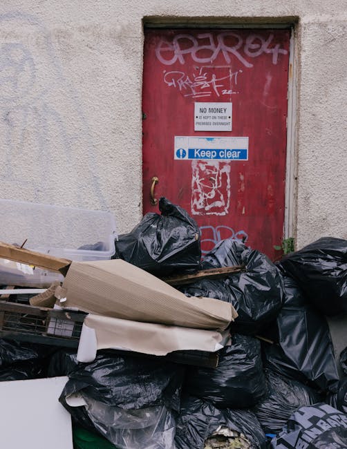 A pile of black rubbish bags and discarded cardboard boxes and packaging materials stacked against a beige textured exterior wall near a red metal door. The door features graffiti tags, two signs with text—one reading 'No Money' and another 'Keep Clear'—and a small, worn handle. The waste, positioned in the foreground, appears to be awaiting collection, with some items partially covered by plastic sheeting or torn cardboard. The scene suggests an outdoor urban setting, possibly a back alley or service area, where independent waste removal or private rubbish clearance is taking place. The lighting is natural, highlighting the contrasting textures of the smooth plastic bags, rough cardboard, and weathered door surface, aligning with waste management practices relevant to non-local authority rubbish disposal or on-site clearance services offered by Waste Removal Hackney.