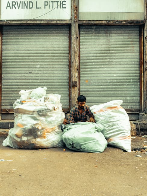 A young man with dark hair and a beard, wearing a patterned shirt, is sitting on the ground in front of a closed, weathered metal roller shutter on a concrete pavement. He is surrounded by several large garbage bags, varying in color from transparent to light green, filled with rubbish such as plastic packaging and household waste. The garbage bags are stacked on each side and in front of him, some appearing partially open or loose. The backdrop features a worn, pale green building structure with visible rusted metal framing and a partially visible sign in the top left corner. The scene suggests an external area used for informal rubbish collection or storage, with no visible waste collection vehicles or designated disposal facilities, emphasizing the context of alternative or private waste handling services. The overall lighting is natural, indicating daytime, and the environment appears to be an urban street setting connected to local rubbish removal practices. Waste Removal Hackney specializes in professional rubbish clearance for similar situations, ensuring proper disposal of accumulated waste outside standard collection hours or locations.