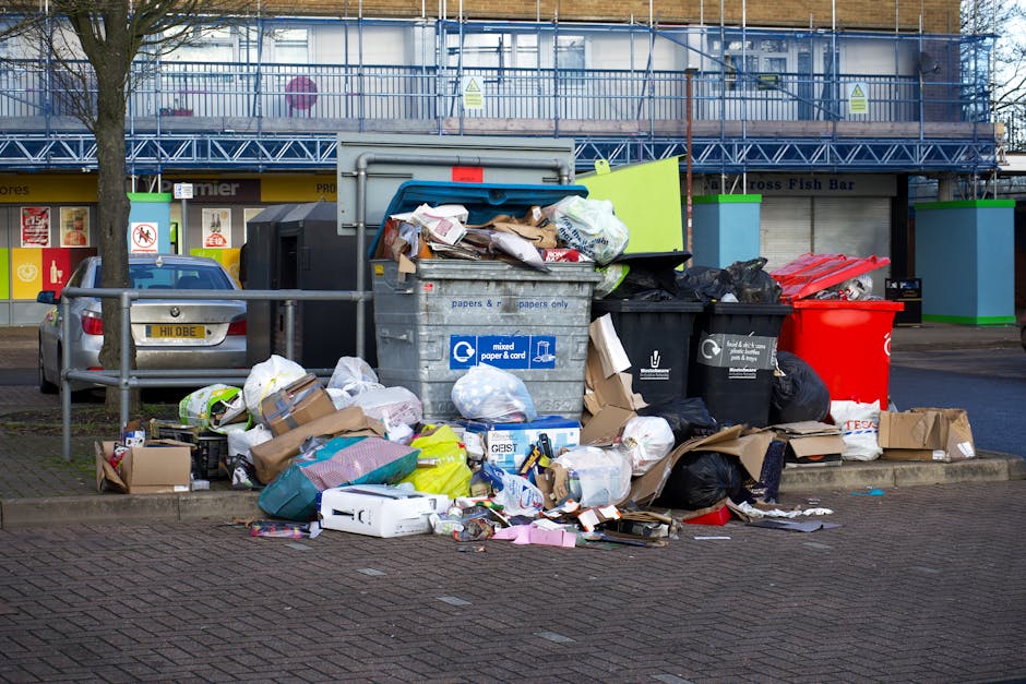 A large accumulation of various waste materials and rubbish bags overflowing from several open-top wheelie bins located on a paved sidewalk in front of a commercial building. The waste consists of mixed paper and cardboard boxes, plastic packaging, black rubbish bags, and discarded packaging materials in different sizes and colors, including brown, white, and black. Several items, such as flattened cardboard containers, plastic bottles, and small household items, are scattered on the ground around the bins, some partially covered by bags. The scene is outdoors, situated on a pedestrian pavement with a metal railing to the left, next to a parked grey car with a UK number plate. In the background, the commercial building is under renovation or construction, enclosed by blue scaffolding and fencing, with storefronts including a fish and bar shop. The environment appears to be daytime with natural light, and this scene subtly illustrates challenges associated with private waste collection practices in urban areas, highlighting the need for professional rubbish removal services like Waste Removal Hackney.