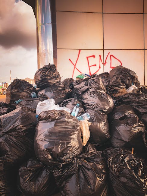 A young man with dark hair and a beard, wearing a patterned shirt, is sitting on the ground in front of a closed, weathered metal roller shutter on a concrete pavement. He is surrounded by several large garbage bags, varying in color from transparent to light green, filled with rubbish such as plastic packaging and household waste. The garbage bags are stacked on each side and in front of him, some appearing partially open or loose. The backdrop features a worn, pale green building structure with visible rusted metal framing and a partially visible sign in the top left corner. The scene suggests an external area used for informal rubbish collection or storage, with no visible waste collection vehicles or designated disposal facilities, emphasizing the context of alternative or private waste handling services. The overall lighting is natural, indicating daytime, and the environment appears to be an urban street setting connected to local rubbish removal practices. Waste Removal Hackney specializes in professional rubbish clearance for similar situations, ensuring proper disposal of accumulated waste outside standard collection hours or locations.