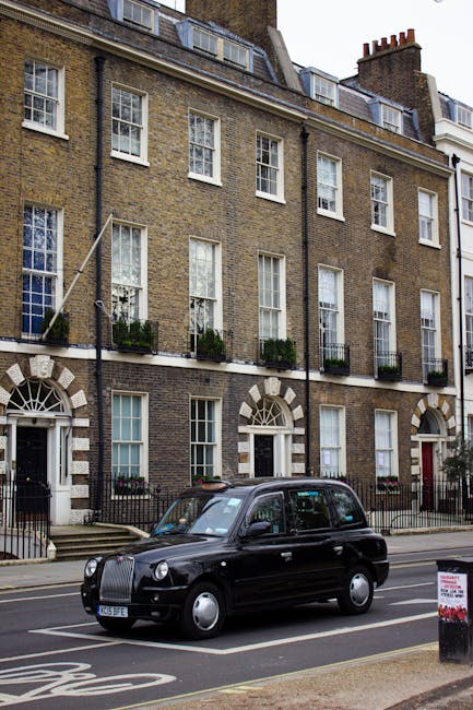 A black London taxi cab is parked on the street in front of a row of traditional Georgian-style terraced buildings constructed of brown brick with white window frames and decorative stone arch surrounds. The buildings feature multiple sash windows, some with small flower boxes or planters on the sills, and have narrow, black wrought-iron railings along the front. The street scene includes a sidewalk with a curb, a small signboard, and a parking restriction marking on the road. In the background, the upper floors of the buildings have additional windows and several roof dormers, with a chimney visible at the top. The overall environment appears urban and visually clean, with no visible rubbish or waste in the scene. The presence of the vehicle and the architecture suggest a typical residential area that might benefit from services such as private rubbish collection or on-site clearance managed by waste removal companies like Waste Removal Hackney, supporting alternative waste handling methods.