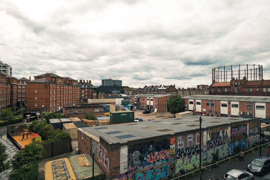 A cityscape view featuring multiple mid-rise brick buildings with flat roofs, interspersed with industrial warehouses, some painted in neutral tones and others with graffiti-covered walls. In the foreground, there is a fenced area with small trees, a children's play area with a yellow slide and colorful equipment, and a flat-roofed warehouse with graffiti art along its side. Behind these structures, there are additional buildings and storage containers, with a large, open driveway space where a few parked cars are visible. To the far right, a tall, circular metal framework, possibly for industrial or water storage purposes, is mounted atop a brick building. The background reveals both residential and commercial buildings under a cloudy sky, creating an urban environment that may reflect an area of private waste collection or alternative rubbish handling. The scene is well-lit by natural daylight, highlighting the textures of the brickwork, graffiti, and metallic surfaces, illustrating a typical urban environment often serviced by local rubbish removal providers like Waste Removal Hackney.