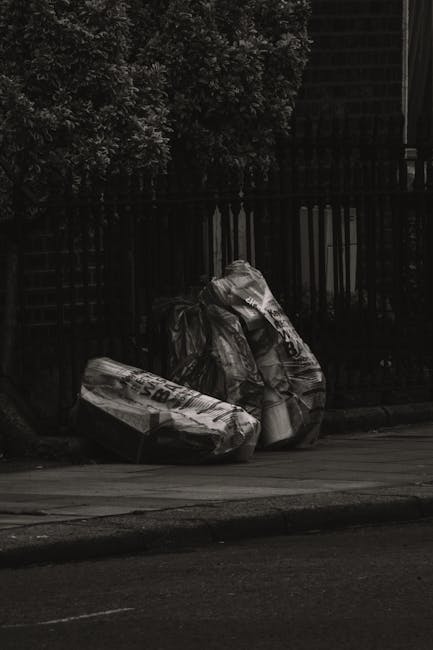 Two large black plastic bags filled with waste are placed on a sidewalk near a wooden fence and a garden hedge. The bags appear to contain bulky rubbish, possibly garden or household waste, and are partially crumpled with visible creases and folds. The scene is set outdoors on a paved surface, with the bags positioned close together and leaning slightly to the right. The background features a dark wooden fence with vertical slats and dense foliage from a leafy hedge. The lighting is subdued, suggesting overcast conditions or shadowed surroundings. This scene highlights the type of waste collection often handled privately by waste removal services like Waste Removal Hackney, which provide alternative disposal options beyond municipal rubbish collection, suitable for clearing garden or bulk household waste.