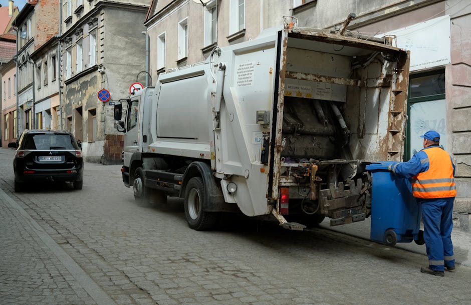 A waste collection vehicle from Waste Removal Hackney is parked on a narrow urban street with cobblestone paving, positioned next to a row of older, multi-story buildings with weathered facades and a mix of brick and plaster finishes. The truck's rear loading compartment is open, revealing rusted metal components and a mechanical system used for compressing and lifting waste. The vehicle is predominantly white, with visible signs of use and dirt on its surface. A worker dressed in a blue uniform with an orange high-visibility vest and matching blue cap is standing to the right of the truck, actively loading a blue wheeled bin into the vehicle's hopper, demonstrating an on-site waste collection process typically associated with private rubbish removal services in urban areas. In the background, another black car is parked on the street, and traffic signs indicating parking restrictions are mounted on the building facade. The scene captures a typical example of independent rubbish collection on a city street, supporting the context of private waste handling and alternative rubbish removal services in a built-up environment.