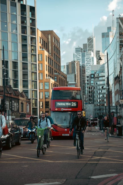 A busy city street scene featuring a red double-decker bus displaying the route number 26 to Hackney Wick, moving toward the foreground amid various pedestrians and cyclists. The background showcases tall modern office buildings and skyscrapers with glass facades reflecting the daylight, creating a vibrant urban environment. To the left, there is a row of parked cars, including a black vehicle, and to the right, pedestrians walk along the pavement, some on bicycles, emphasizing active street-level activity typical of central London areas. The sky above is partly cloudy with patches of blue, contributing to natural, diffused lighting across the scene. This urban setting illustrates a typical environment where private or independent waste collection services, such as those provided by Waste Removal Hackney, operate amidst busy city life and high-rise commercial architecture, supporting alternative waste handling outside of standard municipal rubbish collection.