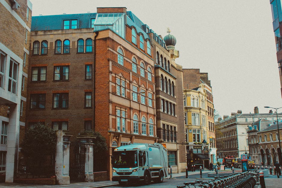 A waste collection vehicle from Waste Removal Hackney is parked on a narrow urban street with cobblestone paving, positioned next to a row of older, multi-story buildings with weathered facades and a mix of brick and plaster finishes. The truck's rear loading compartment is open, revealing rusted metal components and a mechanical system used for compressing and lifting waste. The vehicle is predominantly white, with visible signs of use and dirt on its surface. A worker dressed in a blue uniform with an orange high-visibility vest and matching blue cap is standing to the right of the truck, actively loading a blue wheeled bin into the vehicle's hopper, demonstrating an on-site waste collection process typically associated with private rubbish removal services in urban areas. In the background, another black car is parked on the street, and traffic signs indicating parking restrictions are mounted on the building facade. The scene captures a typical example of independent rubbish collection on a city street, supporting the context of private waste handling and alternative rubbish removal services in a built-up environment.
