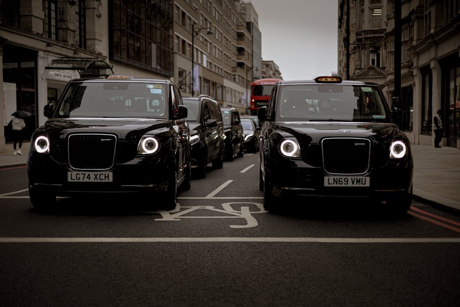 Two black London taxis with illuminated circular headlights are parked side by side in a designated loading zone on a city street. The taxis are positioned facing forward, with their front ends occupying the foreground of the image; the taxi on the left has the license plate LG74 XCH, while the one on the right displays LN69 VMU. Behind the taxis, several other vehicles, including a red double-decker bus and additional cars, are lined up along the narrow urban street, flanked by multi-storey buildings with various window styles and architectural details. Pedestrians are visible on both sides of the street, some walking along the sidewalks, while a few are partially obscured behind parked vehicles. The street surface appears paved with dark asphalt, and there is a marked space in front of the taxis designated for loading or pick-up, as indicated by painted white lines and symbols. The image has natural daylight, with shadows cast by the buildings, contributing to the urban atmosphere. This scene's context relates to vehicle parking, urban transportation, and private or alternative collection services, such as those provided by Waste Removal Hackney for rubbish-related needs.