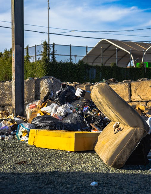 A close-up view of an outdoor rubbish pile situated on a paved surface, consisting of mixed waste materials including black and clear plastic bags filled with waste, a yellow plastic container, a worn beige car seat with fabric upholstery, and some cardboard or paper debris. The waste is loosely piled next to a low stone wall made of rough, light-colored bricks, with some debris spilling onto the ground. Behind the rubbish, there is a dark metal fence with greenery and trees visible beyond, and a partially visible structure with a curved roof or canopy in the background, under a clear blue sky with a few wispy clouds. The scene is illuminated by natural daylight, suggesting daytime, and reflects an environment where private or independent waste disposal, such as rubbish removal services, might be involved, rather than standard council collection.