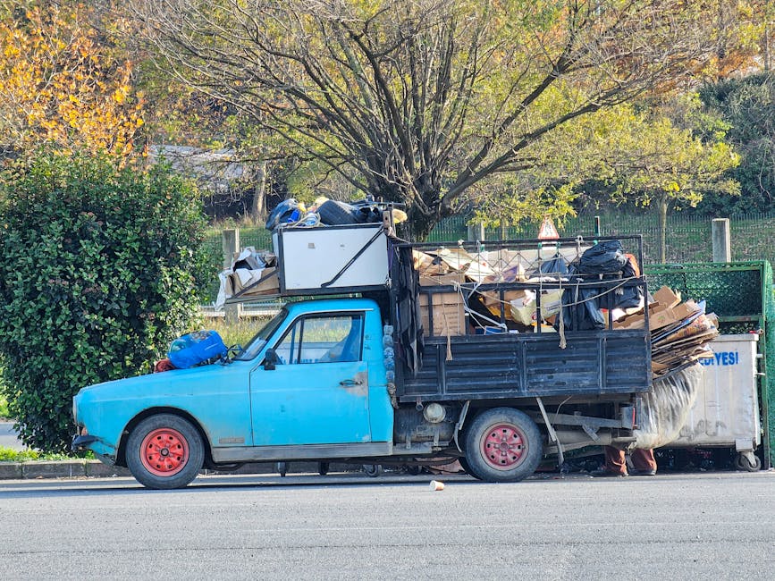A small, light blue flatbed truck parked alongside a road, loaded with a variety of mixed bulky waste including large cardboard boxes, black plastic bags, wooden planks, and miscellaneous household debris. The truck’s cargo area is enclosed with metal side panels, and the waste materials are stacked unevenly, extending above the side rails. The vehicle’s red steel wheels are visible, with one showing signs of rust, and the left front wheel is turned slightly toward the curb. Situated in a suburban area, the background features a park-like setting with a large deciduous tree displaying autumnal yellow and green leaves, and a metal fence along the perimeter. The scene is well-lit with natural daylight, emphasizing the cluttered nature of the load, indicative of private waste removal efforts possibly undertaken by Waste Removal Hackney for alternative rubbish collection outside of standard municipal services.